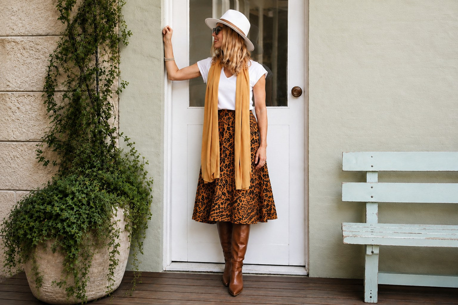 A woman stands on a porch wearing a white hat, white top, tan scarf, brown leopard-print skirt, and brown knee-high boots, posing in front of a white door with a potted plant and bench nearby.